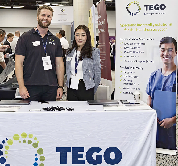 Two individuals standing behind a table at a conference. The table, with a Tego sign, has promotional materials. They smile, creating a friendly atmosphere.