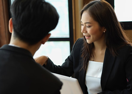 A woman in a business suit smiles while talking to a man, partially visible, both seated at a table with a bright window in the background.