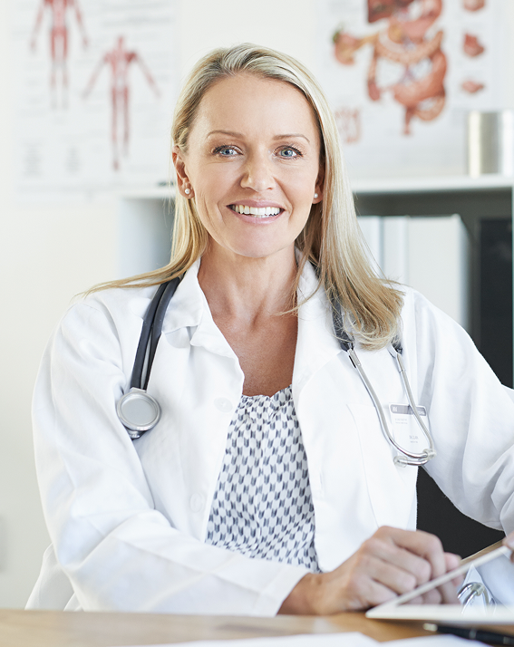 A smiling female doctor sits at a desk, wearing a white coat and stethoscope. Medical charts are visible on the wall, conveying a professional tone.