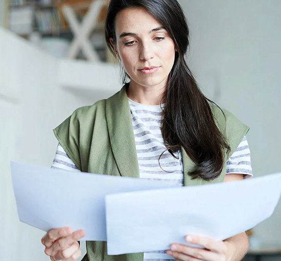 A woman in a striped shirt and green vest looks intently at documents. She appears focused, standing in a bright room with shelves in the background.