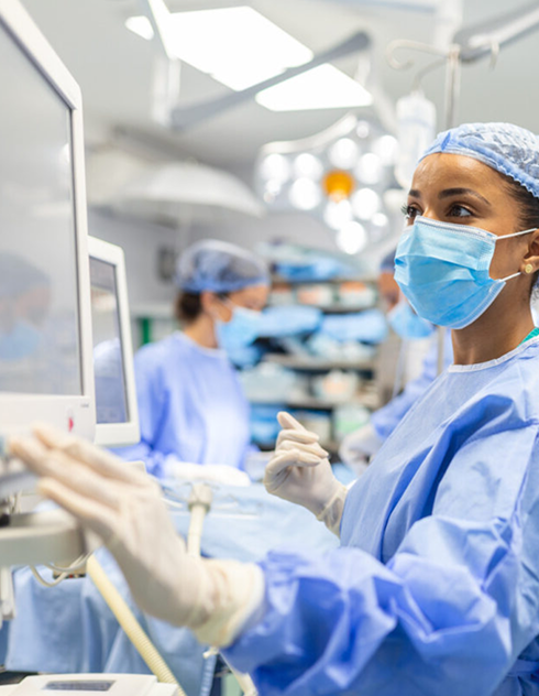 A focused surgeon in blue scrubs and mask operates equipment in a bright, sterile operating room. Another medical professional is seen working in the background.