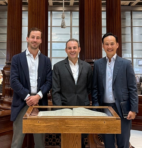 Three men in suits stand smiling behind a display case with an open book inside at Lloyd's of London. The background features large wooden columns and a bright window.