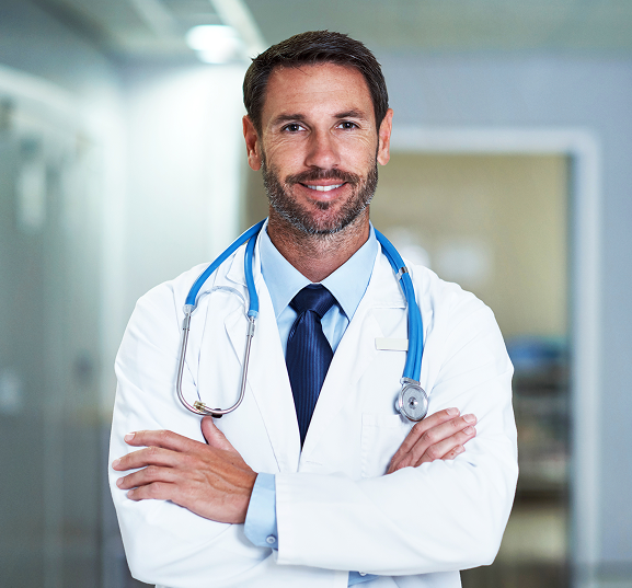 A confident doctor stands with folded arms, smiling warmly. He wears a white coat, blue tie, and stethoscope, set in a bright hospital hallway.