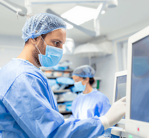 A surgeon in scrubs, mask, and hairnet operates a touchscreen in an operating room. Another masked medical professional works in the background, creating a focused, sterile environment.