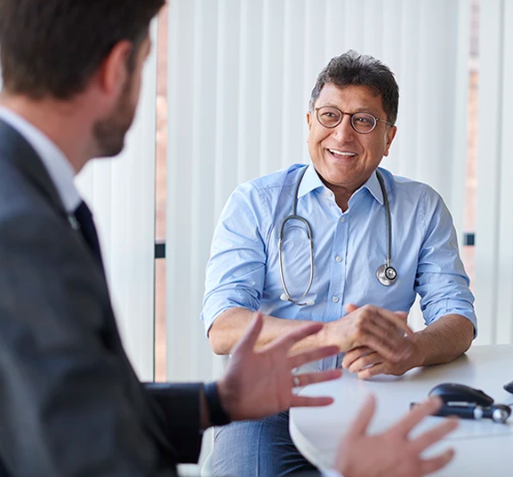 A doctor wearing glasses and a stethoscope smiles engagingly as he talks to a person in a suit inside a well-lit office, creating a friendly atmosphere.
