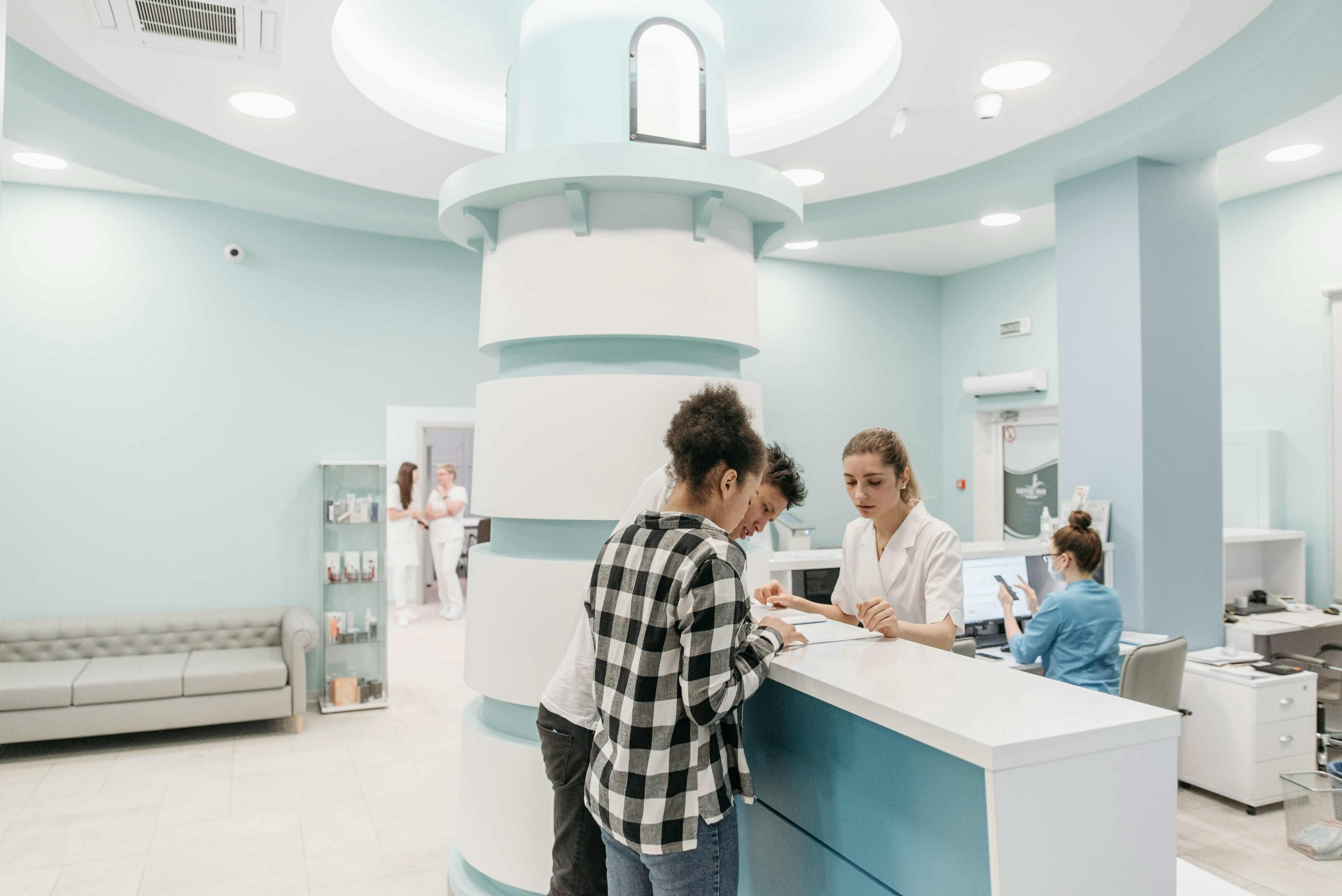 A modern clinic reception with a blue and white colour scheme. Two individuals speak with a receptionist at a large counter. The atmosphere is calm.