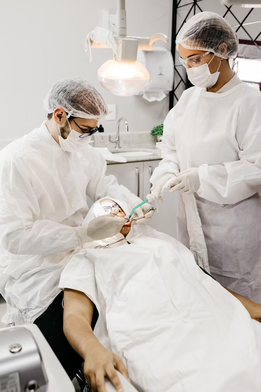 Two dentists in white protective attire and surgical masks work on a patient under a bright examination light in a modern dental clinic.
