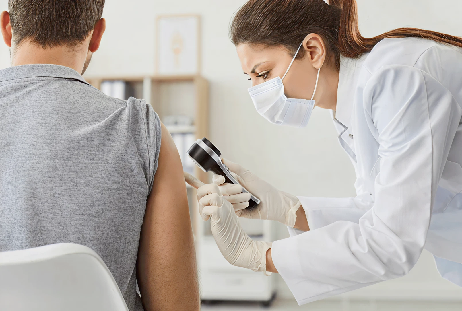A healthcare professional in a mask examines a patient's upper arm with a dermatoscope. The setting is a bright, clinical office. The mood is focused and professional.