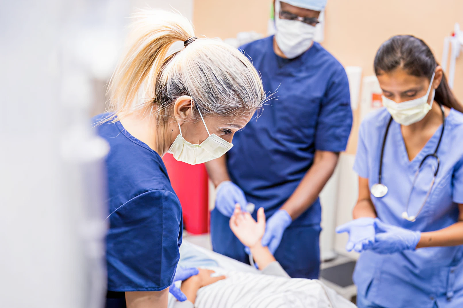 Three healthcare workers in masks and gloves attend to a patient in a hospital setting. They appear focused and attentive, conveying calm professionalism.
