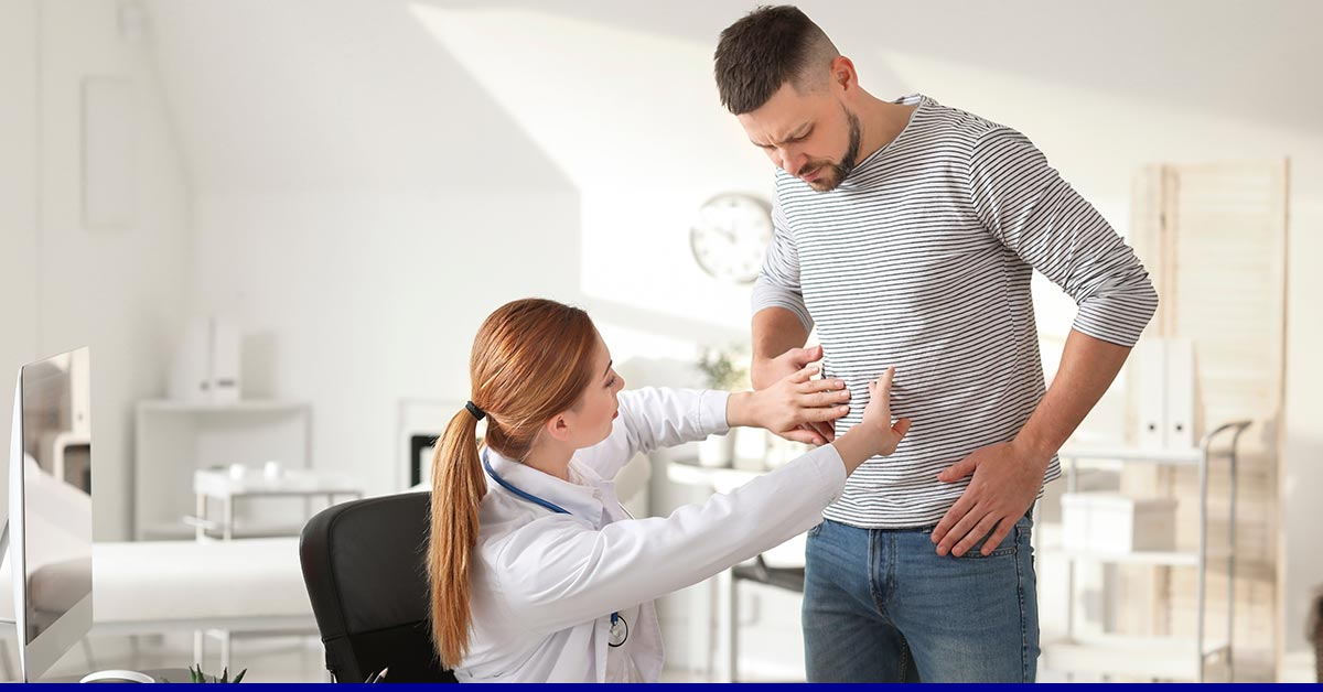 A doctor in a white coat examines a man's abdomen in a bright medical office. The man, in a striped shirt, appears concerned, indicating attention to his discomfort.