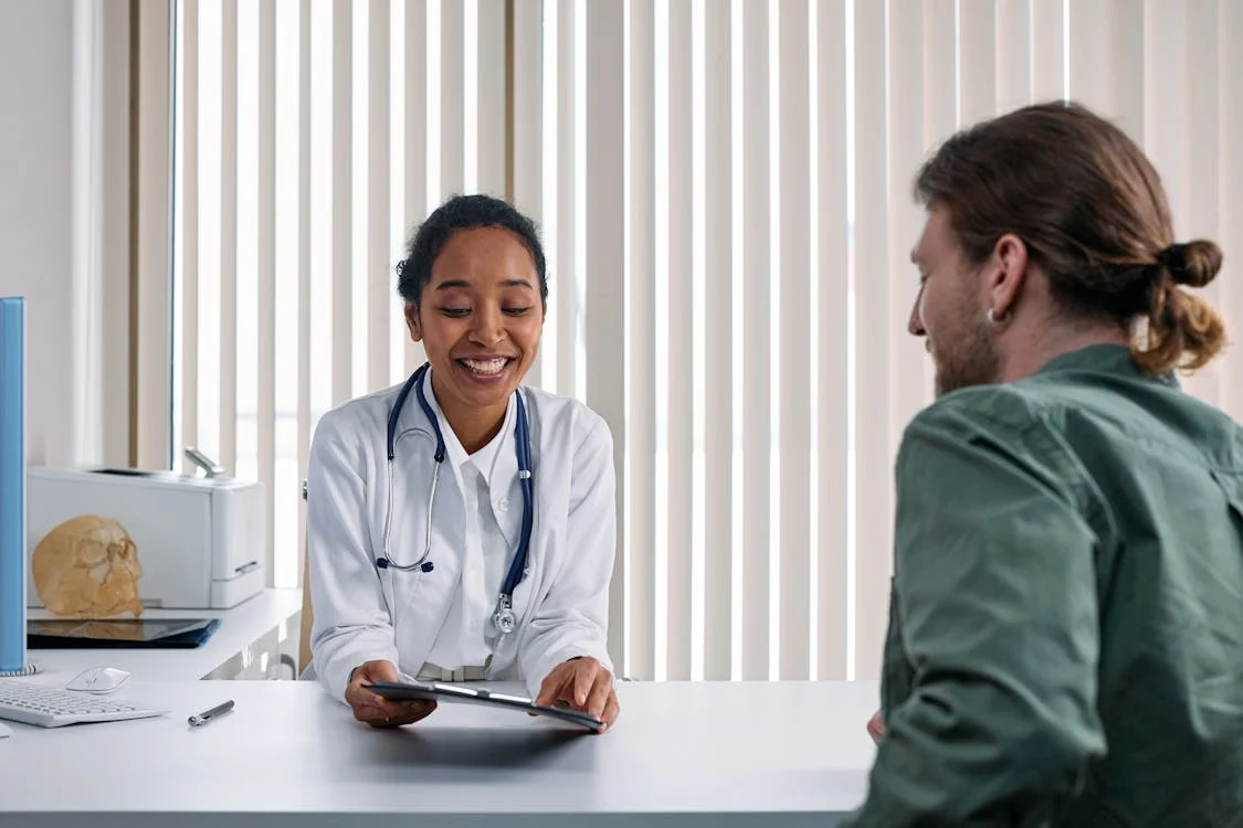 A smiling doctor in a white coat holds a tablet, talking to a patient across a desk. The setting is well-lit with vertical blinds, creating a professional and friendly atmosphere.