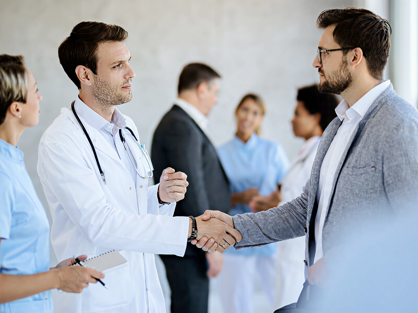 A doctor in a white coat shakes hands with a man in a suit, conveying professionalism. Medical staff and another man converse in the blurred background.
