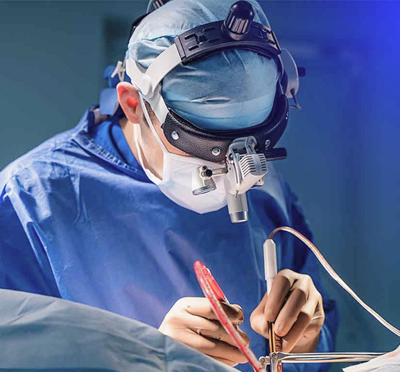 Surgeon in blue scrubs and protective gear focuses intently on a delicate procedure under bright surgical lights, conveying precision and concentration.