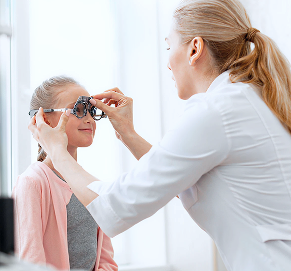 Optometrist adjusting trial frame on child's face during eye exam. The child sits calmly, wearing a pink sweater, in a bright room.