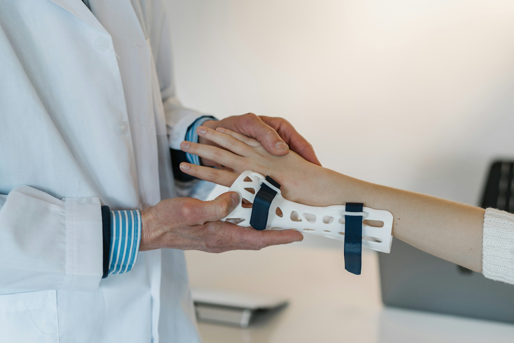 A doctor in a white coat gently examines a patient's arm, which is fitted with a white wrist brace. The setting is calm and clinical.