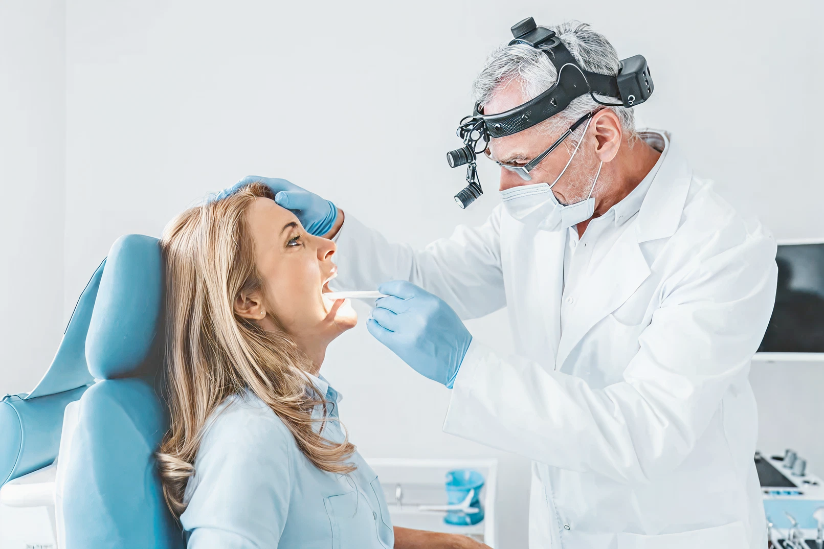 A doctor wearing a surgical mask and headlamp examines a patient's throat with a tongue depressor. The patient is seated, appearing calm. Medical setting.