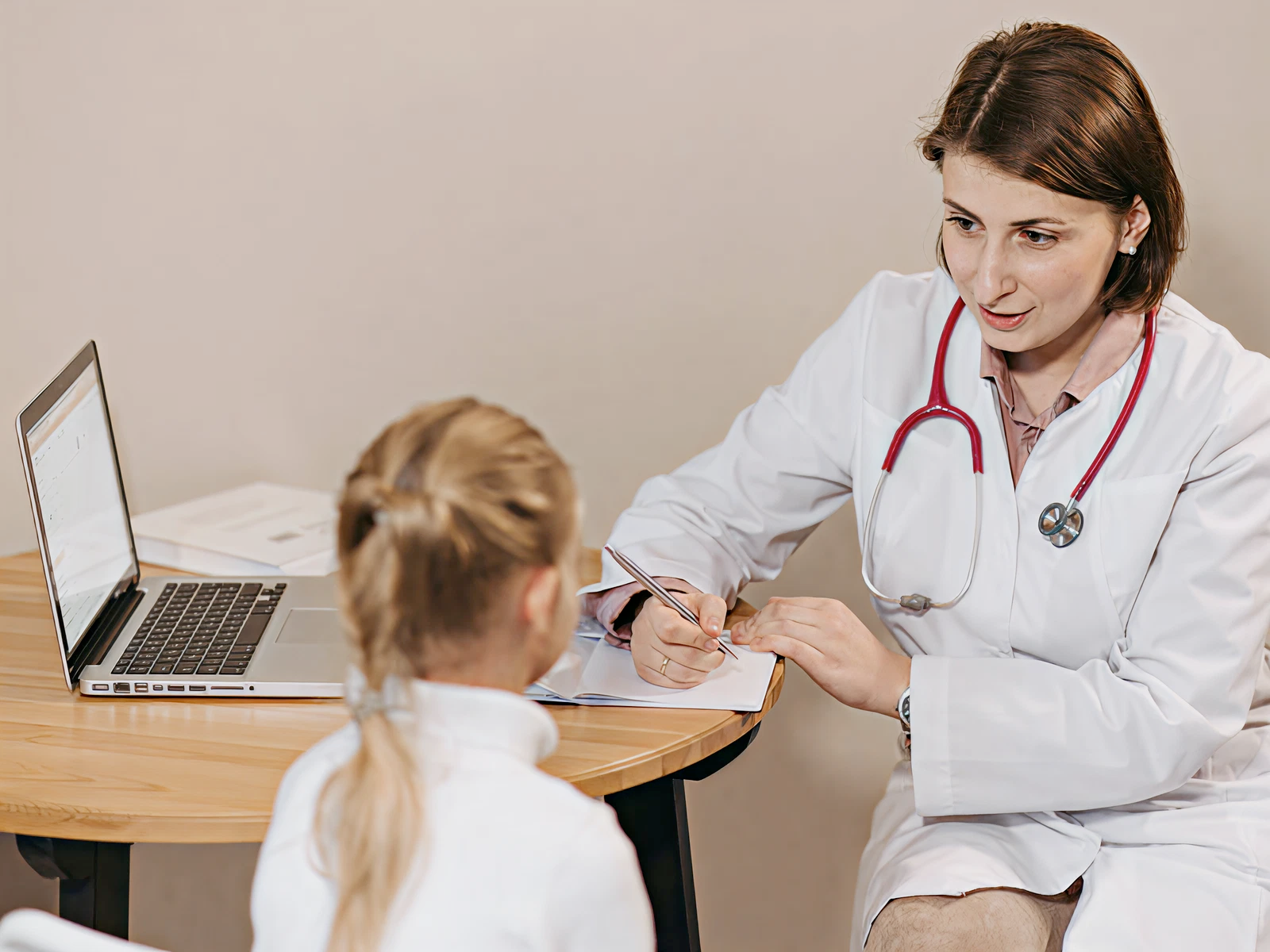 A doctor in a white coat and stethoscope talks to a child at a desk with a laptop. The setting is calm and focused on communication.