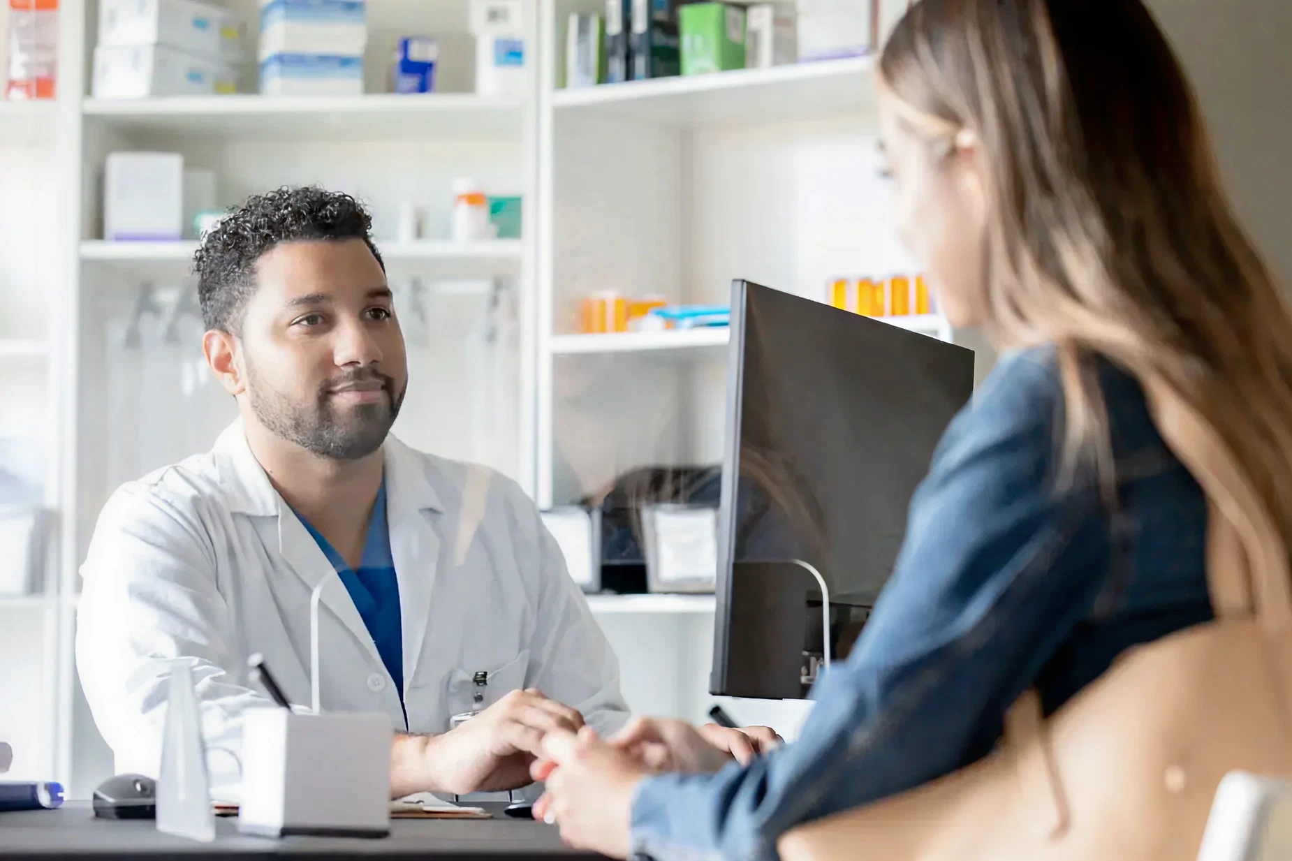 A doctor in a white coat listens attentively to a patient in an office. Shelves with medical supplies are in the background, conveying a professional setting.