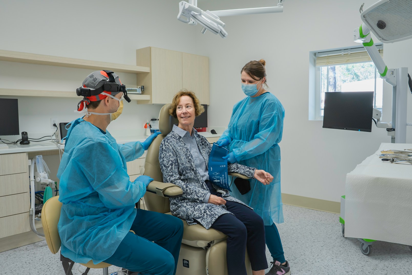 A woman sits in a dental chair, smiling at a dentist in blue scrubs and a visor. A nurse, also in blue scrubs, checks her blood pressure. The setting is a bright, modern clinic.