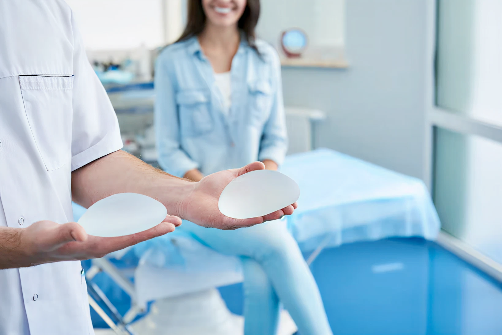 A medical professional holds two breast implants in a clinic, while a smiling woman in casual attire sits on an examination table. The mood is calm and professional.
