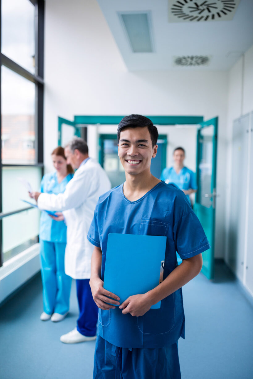 Smiling healthcare worker in blue scrubs holds a clipboard in a hospital hallway. Behind him, colleagues discuss documents, conveying teamwork and care.